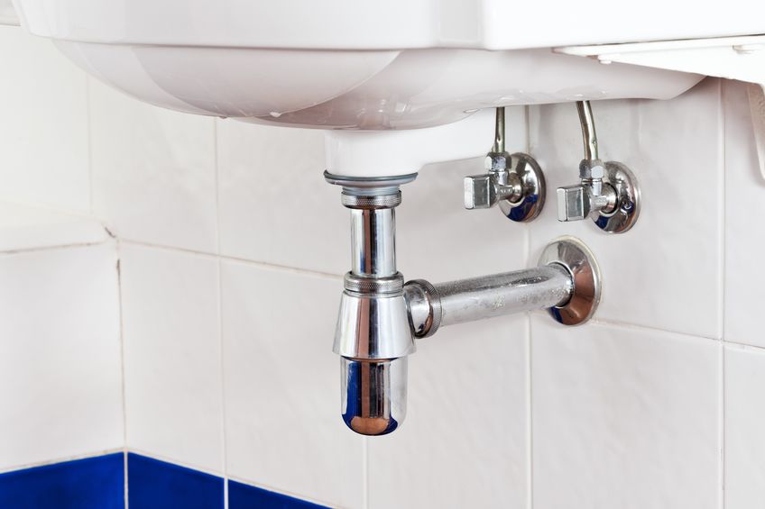 White sink with chrome pipe and two shut-off valves, mounted above white and blue tiled wall.