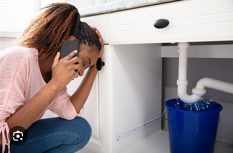 Person on the phone crouching by a leaking pipe under a sink, with water dripping into a blue bucket.