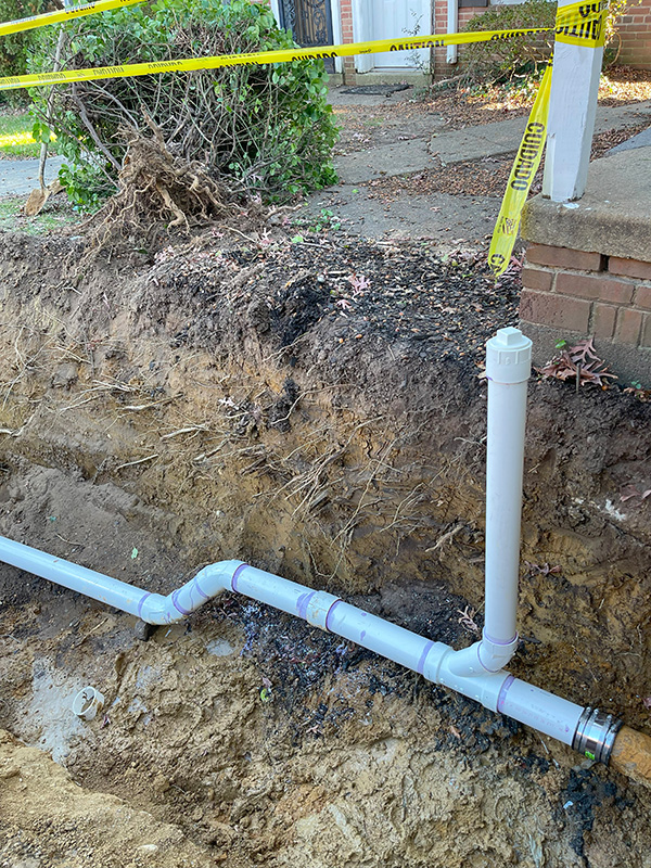 A dug-up trench showing newly installed PVC pipe, with visible fittings, near a brick walkway and yellow caution tape above.