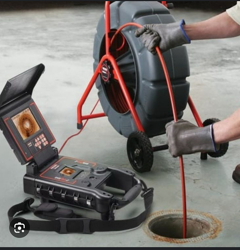 A person wearing gloves uses a plumbing inspection camera to examine a pipe through a hole in a concrete floor.